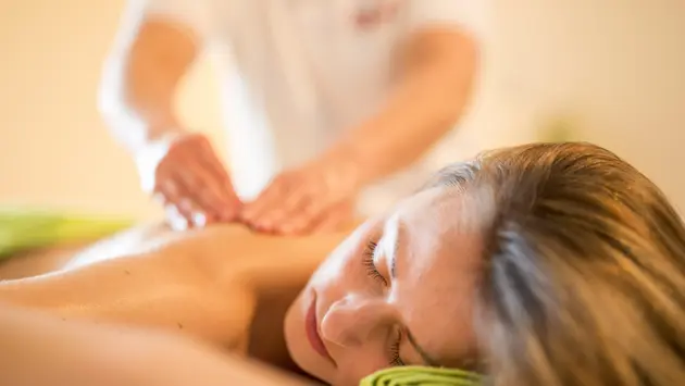 SWG-Massage-Hot-Stone Man sieht eine liegende Frau mit langen Haare, die von einem Masseur am Rücken massiert wird. Die Frau hat ihre Augen geschlossen und genießt die Massage.