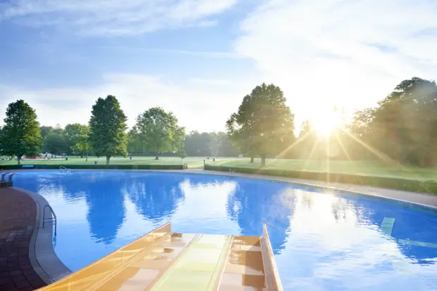 Auf dem Bild erkennt man aus der Perspektive einer Rutsche ein Schwimmbecken im Freibad sowie Grünfläche im Hintergrund und die Sonne, die auf das stille Wasser scheint.