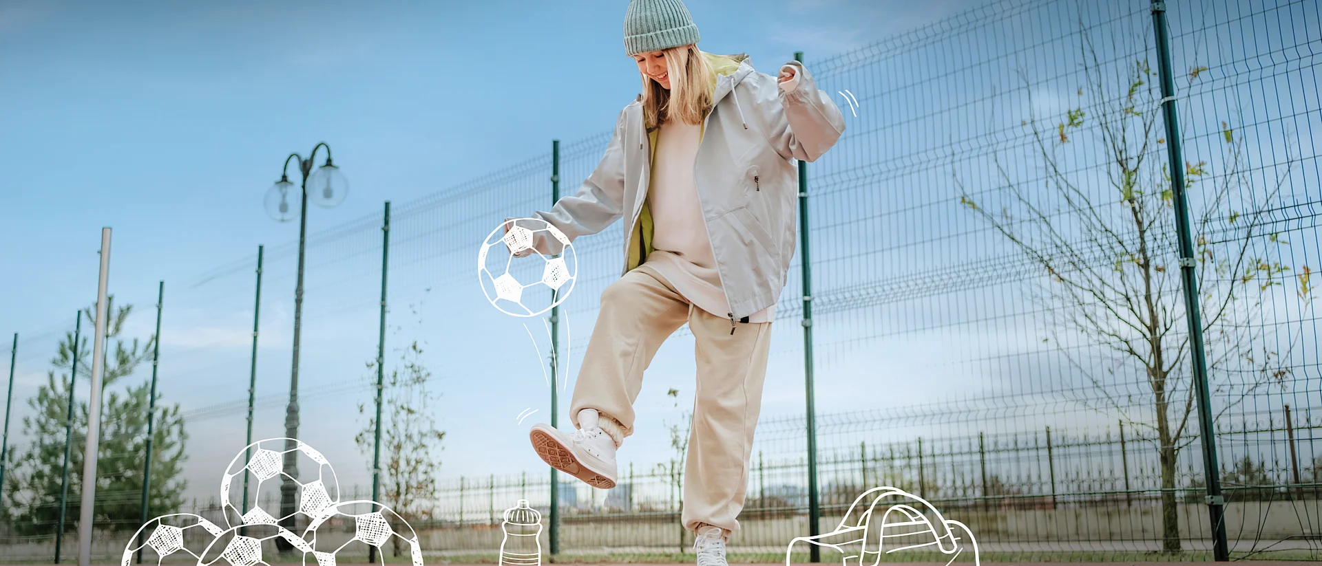 Ein Mädchen in einer hellen Jacke, Jogginghose und Mütze spielt auf einem Sportplatz mit einem Ball. Sie jongliert den Ball mit dem Fuß. Im Hintergrund sind ein hoher Zaun und Bäume zu sehen. Um sie herum sind Zeichnungen von mehreren Fußbällen, einer Trinkflasche und einer Sporttasche auf dem Boden dargestellt.