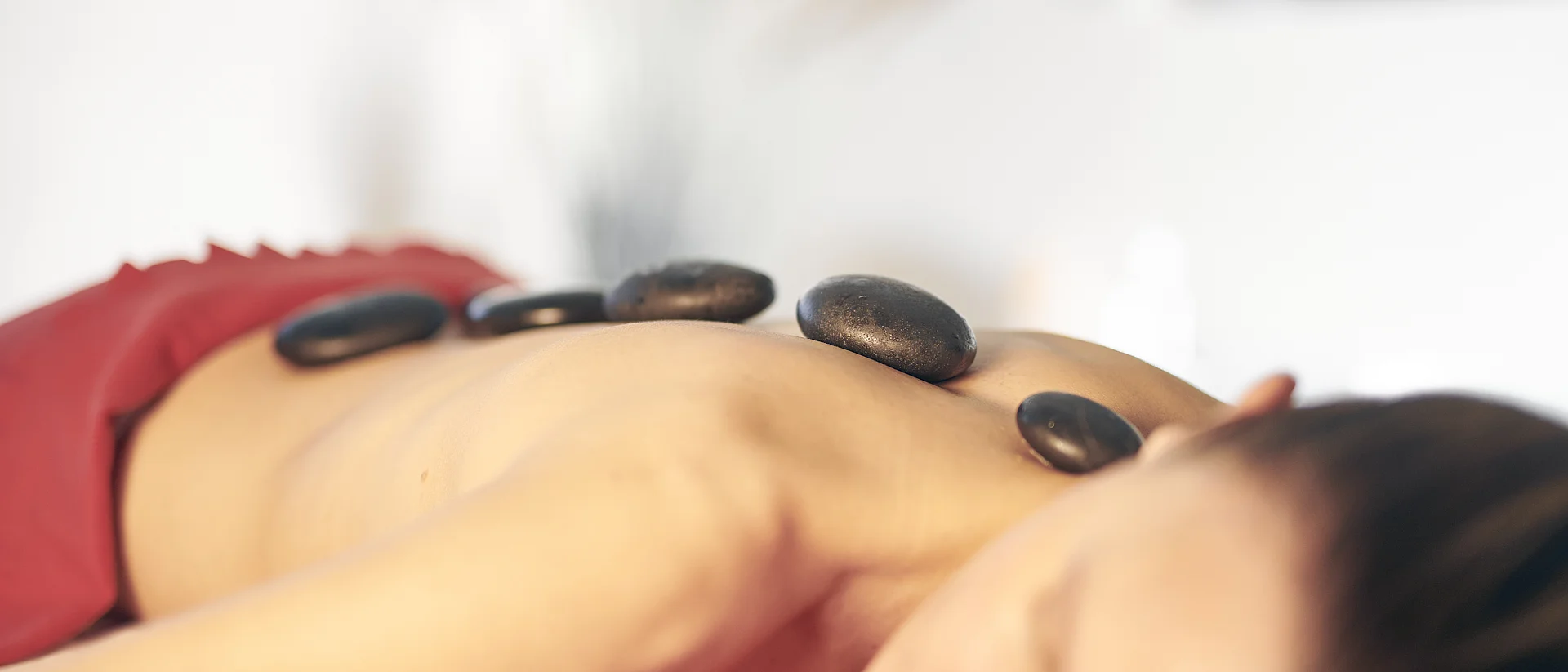 Auf dem Foto erkennt man eine Frau auf einer Massageliege. Ihre Beine sind mit einer Decke überdeckt. Auf ihrem Rücken liegen schwarze heiße Steine.
