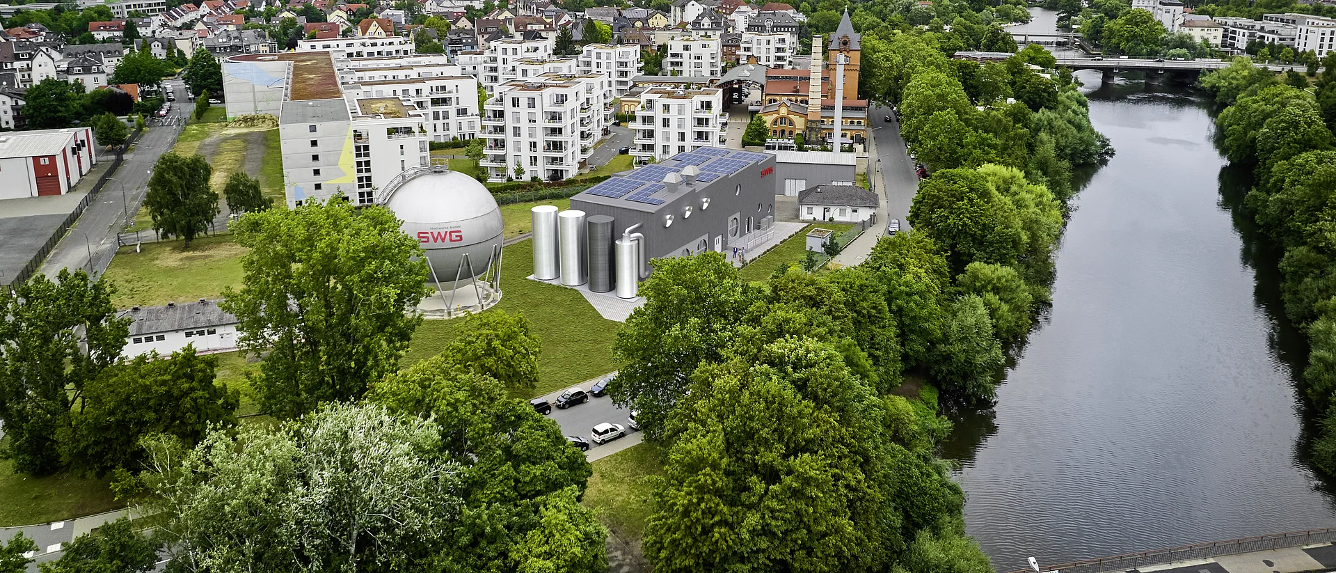 Das Bild zeigt eine Luftaufnahme eines Stadtgebiets mit einem zentralen Gebäudeensemble, bestehend aus einem großen kugelförmigen Gasspeicher mit der Aufschrift „SWG“, mehreren zylindrischen Tanks und einem Flachdachgebäude mit Solarpaneelen. Die Gebäude sind umgeben von Wohnhäusern und viel Grün.