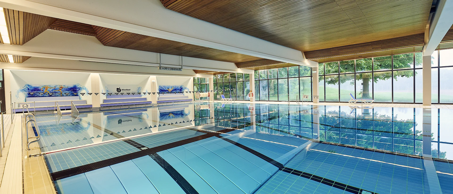 Ein modernes Hallenbad mit einem großen, hellblauen Schwimmbecken, das von Edelstahlgeländern umgeben ist. Die Decke besteht aus Holzpaneelen, die eine warme Atmosphäre schaffen. Im Hintergrund sind große Fenster, die viel Tageslicht hereinlassen und eine grüne Außenansicht bieten.