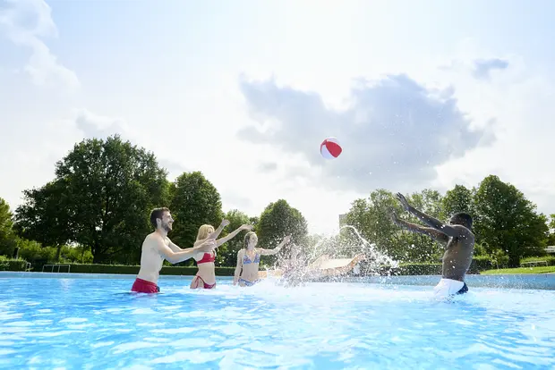 Auf dem Bild sieht man fünf planschende Menschen im Freibad. Am blauen Himmel ist eine Wolke. Der Ball ist in der Luft und das Wasser aus dem Becken spritzt in die Luft.