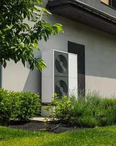 Modern heat pump unit installed next to a residential house wall, surrounded by green lawn, bushes, and young trees in a landscaped yard