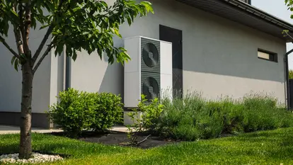 Modern heat pump unit installed next to a residential house wall, surrounded by green lawn, bushes, and young trees in a landscaped yard