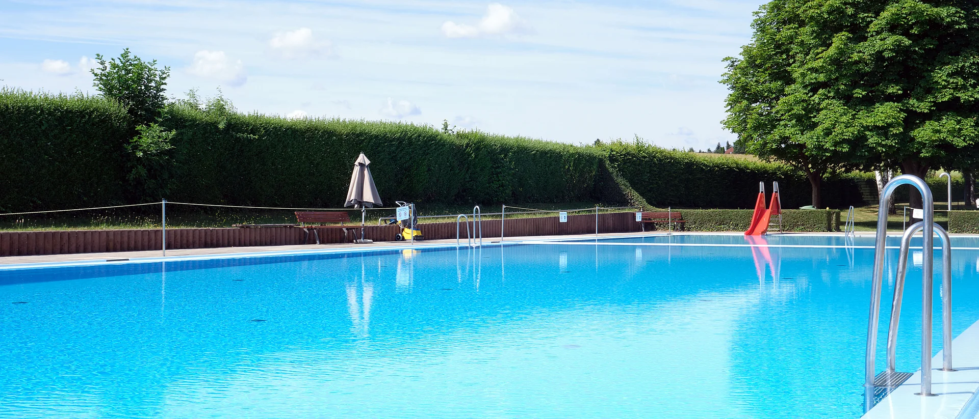 Auf dem Bild sieht man ein ruhiges Freibadbecken mit einem Sonnenschirm und einer kleinen roten Rutsche im Hintergrund. Der Himmel ist blau und auf der rechten Seite sieht man einen großen Baum. 