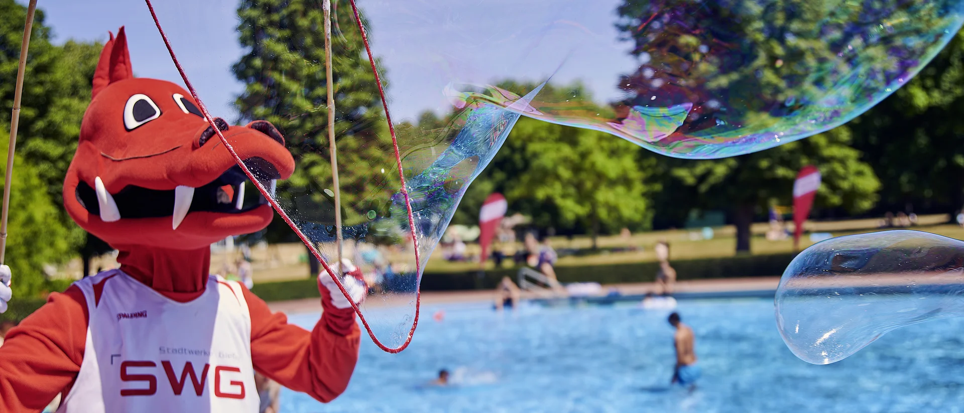 Das Bild zeigt das rote Drachenmaskottchen der Stadtwerke Gießen (SWG) an einem sonnigen Tag im Freien, wahrscheinlich in einem Freibad oder Schwimmbad. Das Maskottchen trägt ein weißes Trikot mit der Aufschrift "SWG" und macht mit einem großen Seifenblasenstab riesige Seifenblasen.