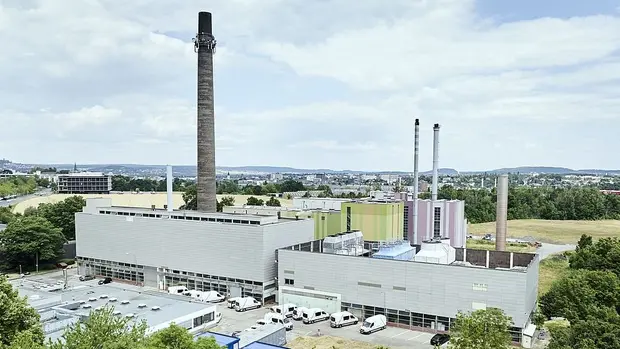 SWG-HKW-Außenaufnahme Das Bild zeigt ein Industriegebäude mit mehreren Schornsteinen, die aus dem Gebäude herausragen. Im Vordergrund ist ein moderner Gebäudekomplex zu sehen, der von mehreren Fahrzeugen umgeben ist, die vor dem Gebäude parken. Im Hintergrund sind einige Bäume und eine offene Fläche zu erkennen, die in die Ferne reicht. Am Horizont ist eine Stadtlandschaft mit weiteren Gebäuden und Hügeln zu sehen.
