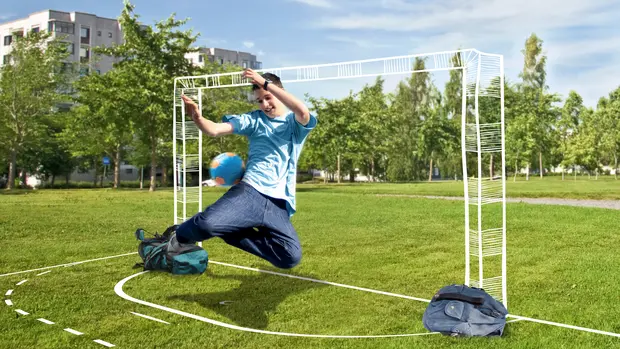 SWG-Spiel-dein-Spiel-Junge-Torhüter Ein Junge in einem hellblauen T-Shirt und Jeans springt mit einem blauen Fußball in der Hand in die Luft, als ob er ein Tor verhindern möchte. Er spielt auf einer grünen Wiese, auf der ein improvisiertes Fußballtor aus zwei Rucksäcken und weißen Linien auf dem Gras markiert ist.