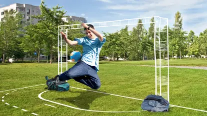 SWG-Spiel-dein-Spiel-Junge-Torhüter Ein Junge in einem hellblauen T-Shirt und Jeans springt mit einem blauen Fußball in der Hand in die Luft, als ob er ein Tor verhindern möchte. Er spielt auf einer grünen Wiese, auf der ein improvisiertes Fußballtor aus zwei Rucksäcken und weißen Linien auf dem Gras markiert ist.