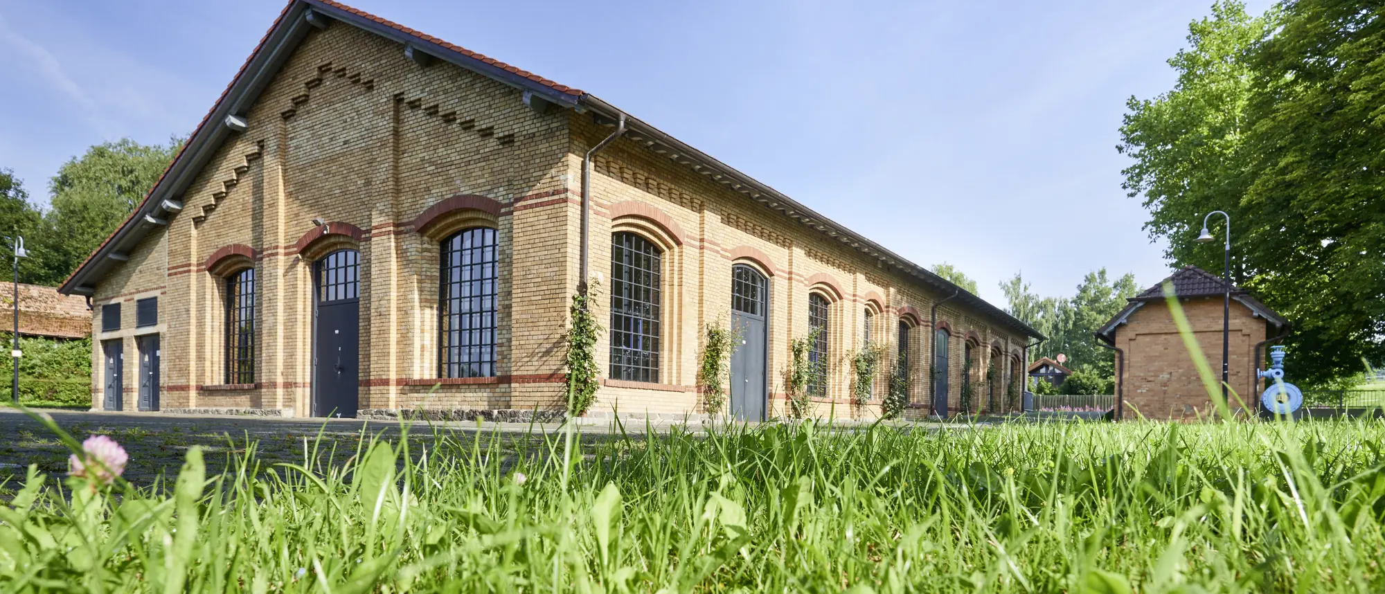 Ein historisches Backsteingebäude des Wasserwerks Queckborn, umgeben von grünen Rasenflächen und Bäumen. Das Gebäude hat große Fenster mit schwarzen Rahmen und steht unter einem klaren, blauen Himmel.