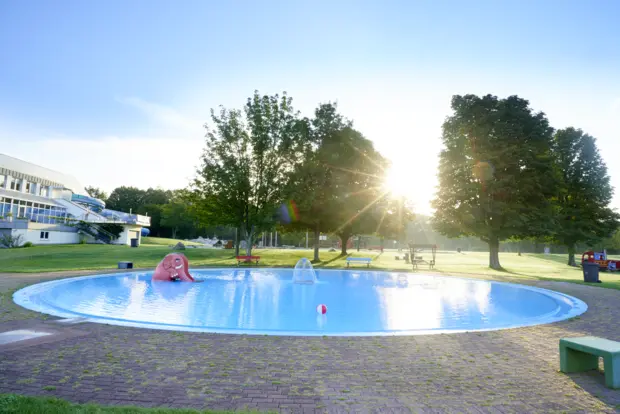 Auf dem Bild erkennt man ein rundes Kinderbecken in einem Freibad mit einer Elefantenrutsche und einem Wasserfall in der Mitte des Beckens. Im Hintergrund befindet sich ein Gebäude mit einer Rutsche sowie Grünfläche und Bäumen.