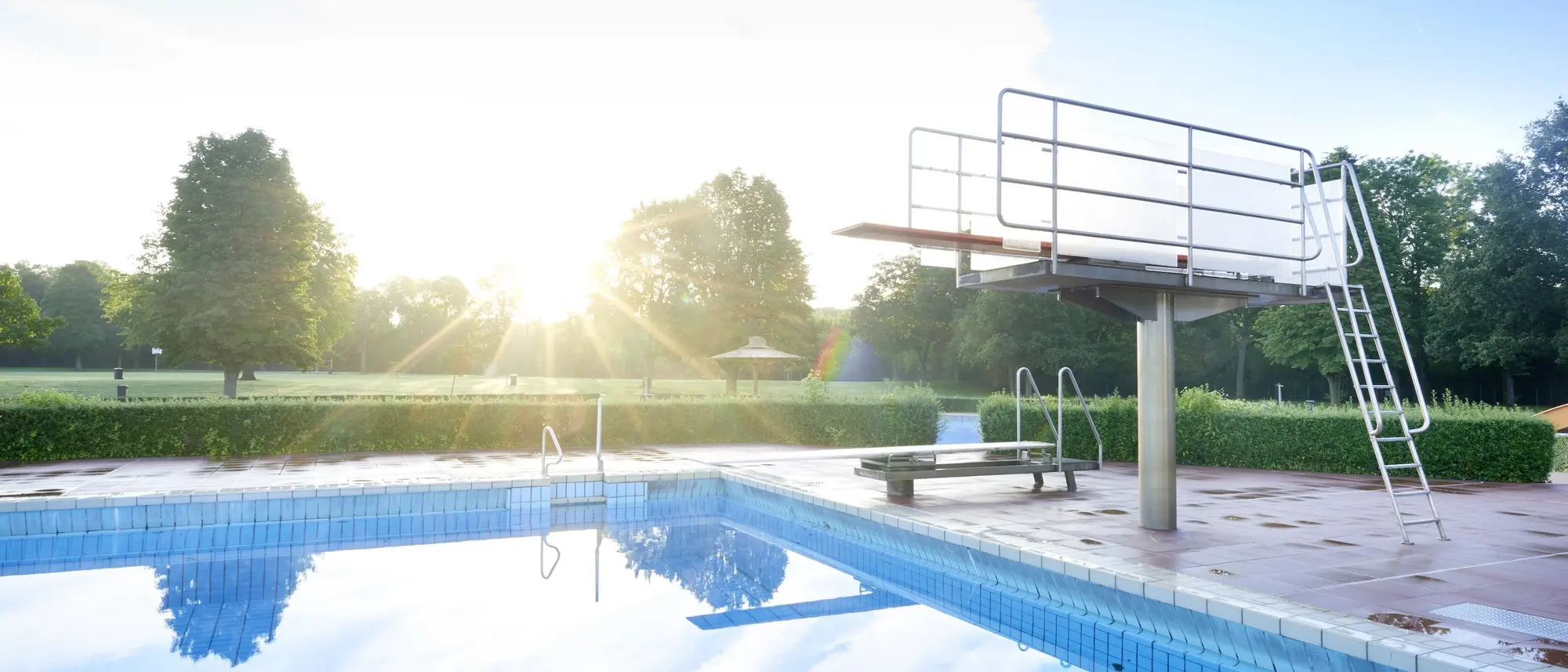 Foto von einem Freibad mit Sprungturm und klaren, stillen Wasser. Die Sonne scheint.