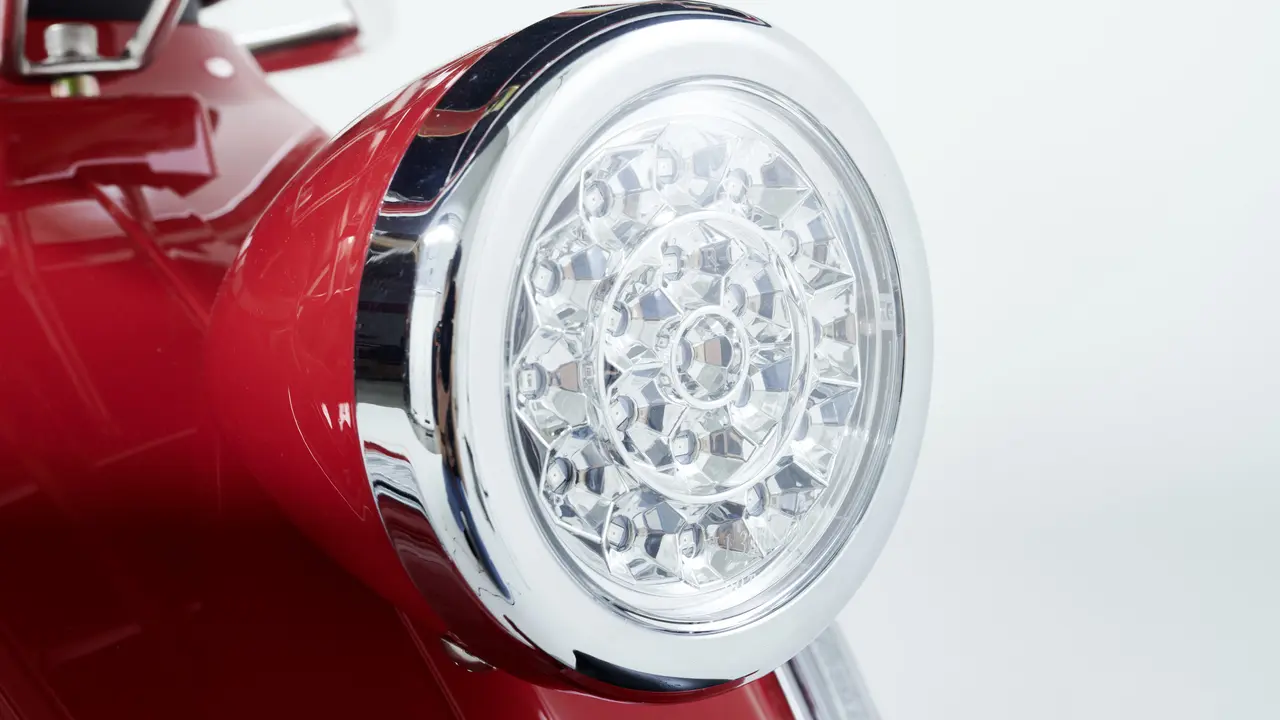  Nahaufnahme eines runden LED-Scheinwerfers eines glänzend roten E-Retro-Rollers. Der Scheinwerfer ist von einem verchromten Rahmen umgeben und zeigt ein detailliertes, facettiertes Design der LED-Leuchten.