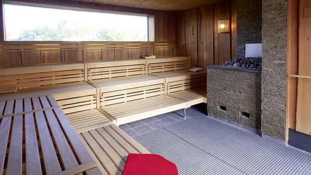 SWG-Sauna-mit Saunaofen Auf dem Foto sieht man eine Sauna mit einem Fenster sowie mit einem Saunaofen auf der rechten Seite. Im Vordergrund liegt ein rotes gefaltetes Handtuch.