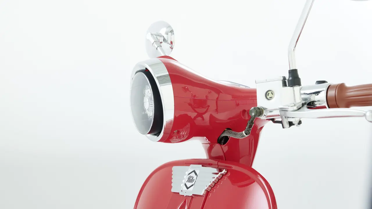  Nahaufnahme des Lenkers und Scheinwerfers eines roten Retro-Rollers, zeigt verchromte Details und den vorderen Teil des Rollers.