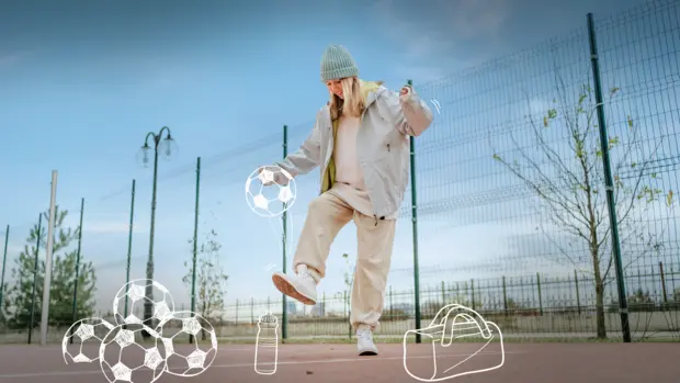 SWG-Spiel-dein-Spiel-Mädchen-Fußball Ein Mädchen in einer hellen Jacke, Jogginghose und Mütze spielt auf einem Sportplatz mit einem Ball. Sie jongliert den Ball mit dem Fuß. Im Hintergrund sind ein hoher Zaun und Bäume zu sehen. Um sie herum sind Zeichnungen von mehreren Fußbällen, einer Trinkflasche und einer Sporttasche auf dem Boden dargestellt.