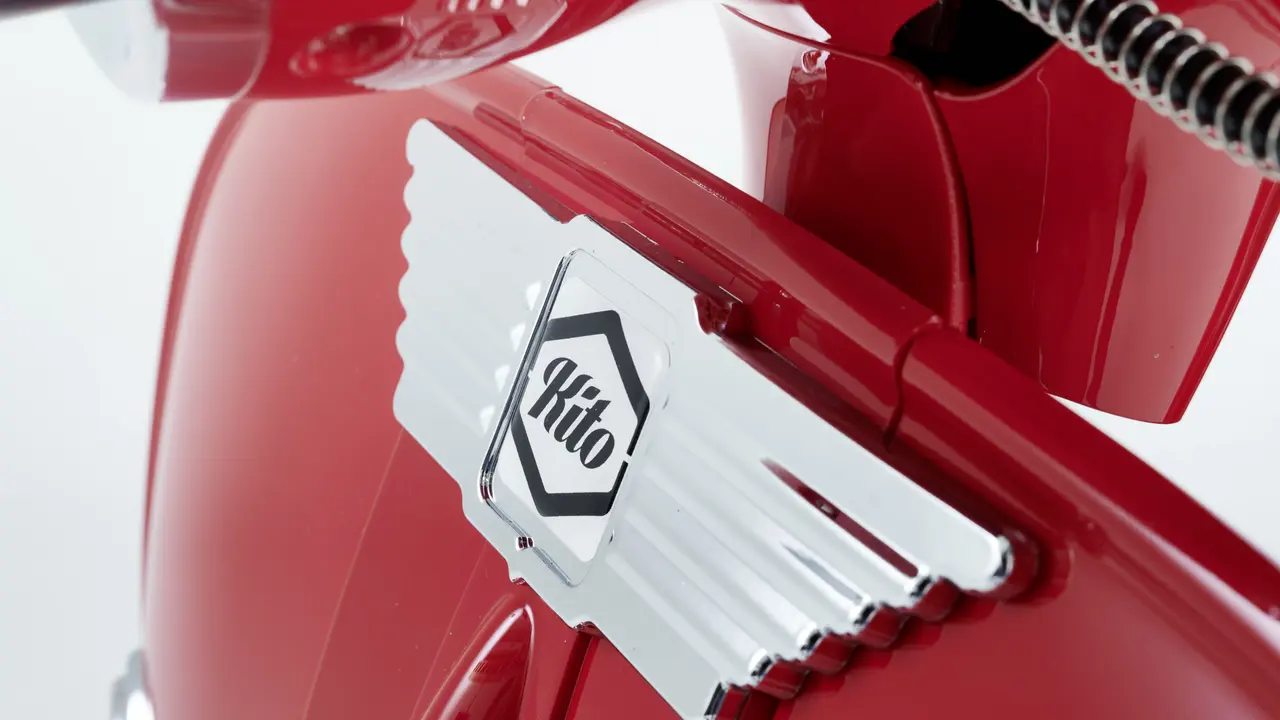 Nahaufnahme des Logos auf dem vorderen Teil eines roten E-Retro-Rollers, zeigt ein verchromtes Emblem mit dem Schriftzug "Kito".