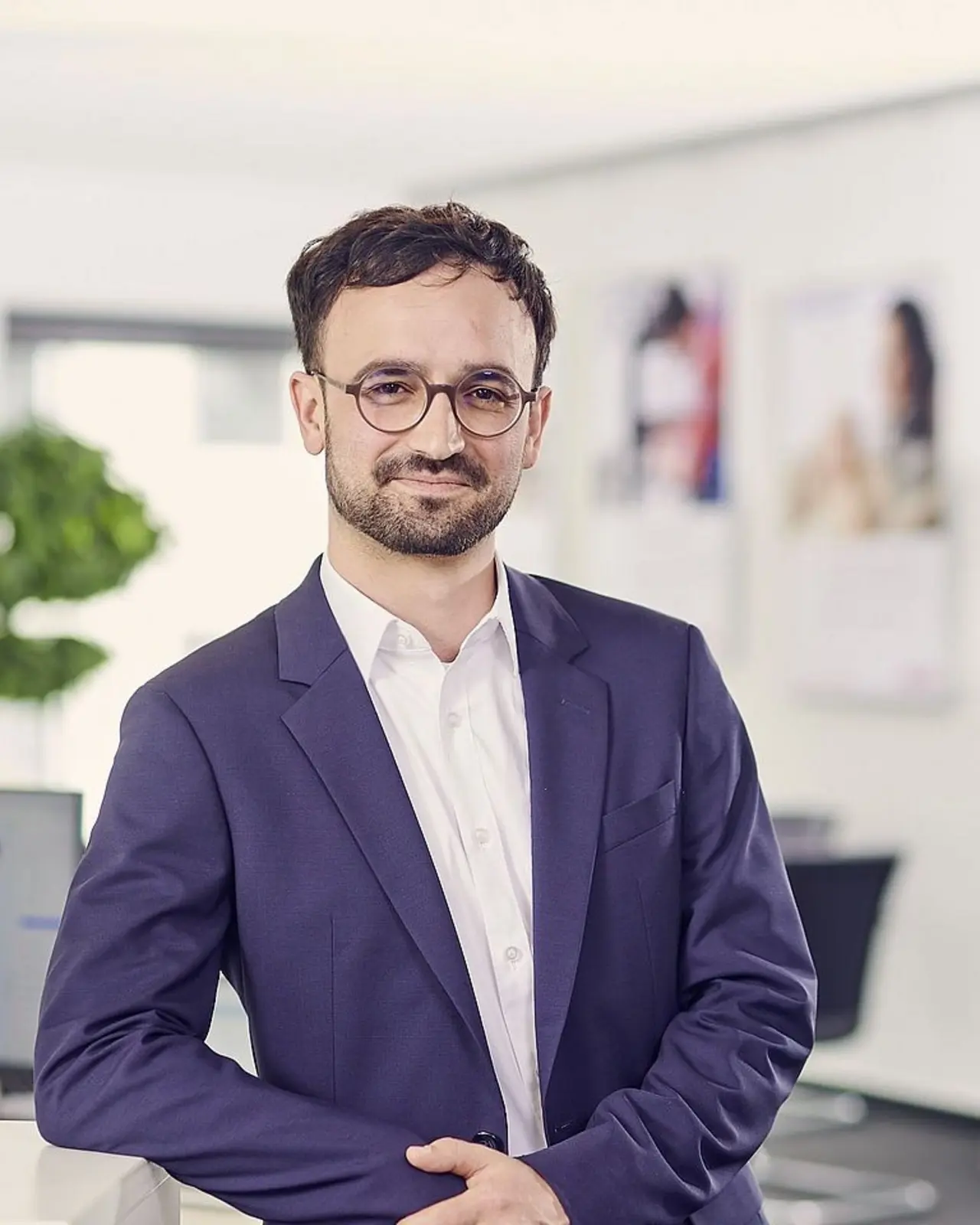 Ein Mann mit Brille und kurzen dunklen Haaren steht in einem modernen Büro. Er trägt einen dunkelblauen Anzug und ein weißes Hemd. Im Hintergrund sind unscharf verschiedene Büroeinrichtungen und Dekorationen zu sehen.