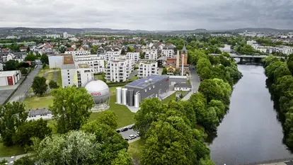 SWG-Flusswasserwärmepumpe Das Bild zeigt eine Luftaufnahme eines Stadtgebiets mit einem zentralen Gebäudeensemble, bestehend aus einem großen kugelförmigen Gasspeicher mit der Aufschrift „SWG“, mehreren zylindrischen Tanks und einem Flachdachgebäude mit Solarpaneelen. Die Gebäude sind umgeben von Wohnhäusern und viel Grün.