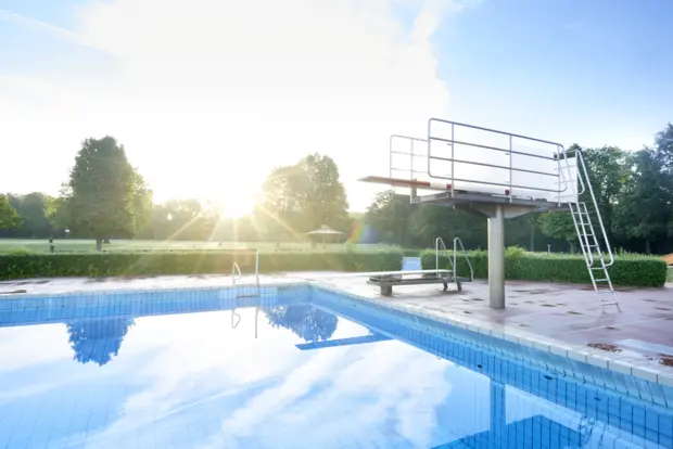 Auf dem Bild erkennt man ein stilles Freibadbecken mit einen hohen und einem niedrigen Sprungturm. Die Sonne strahlt auf das Becken und im Hintergrund sind grüne Bäume und Hecke.