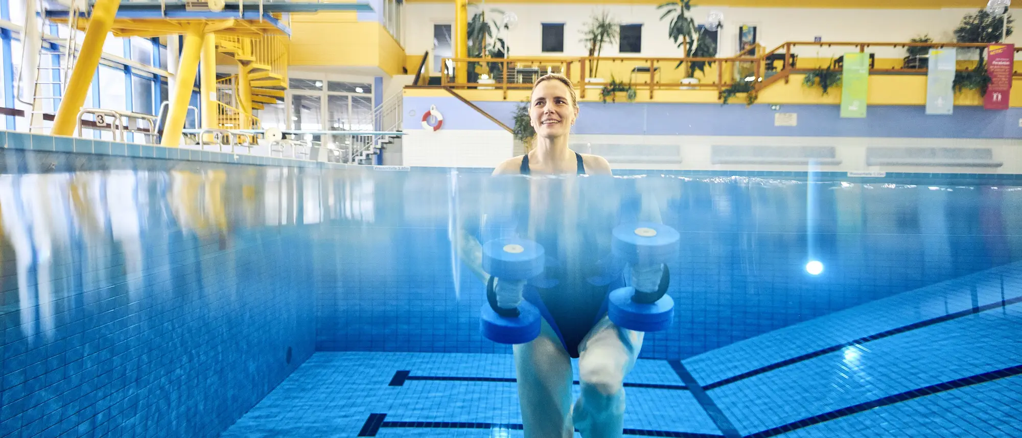 Das Bild zeigt, wie eine Frau unter Wasser Aquajogging betreibt. Dabei sieht man ihren ganzen Körper und wie sie Aquahanteln in der Hand hält.