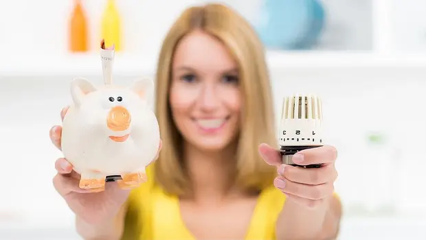 SWG-Sparschwein-Thermostat Das Bild zeigt eine lächelnde Frau, die ein Sparschwein und einen Heizkörperthermostat in die Kamera hält. Das Sparschwein ist weiß mit orangefarbenen Details, und es steckt ein Geldschein aus der Öffnung heraus. Der Heizkörperthermostat, den sie in der anderen Hand hält, ist ähnlich dem, was in Haushalten zur Temperaturregelung verwendet wird.