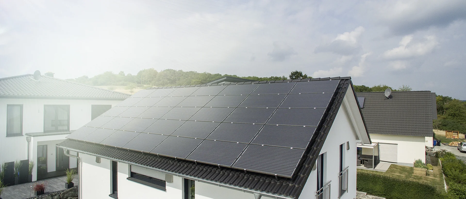  Modernes Einfamilienhaus mit großflächigen Solarpanelen auf dem Dach, sonniger Tag, benachbarte Häuser im Hintergrund.