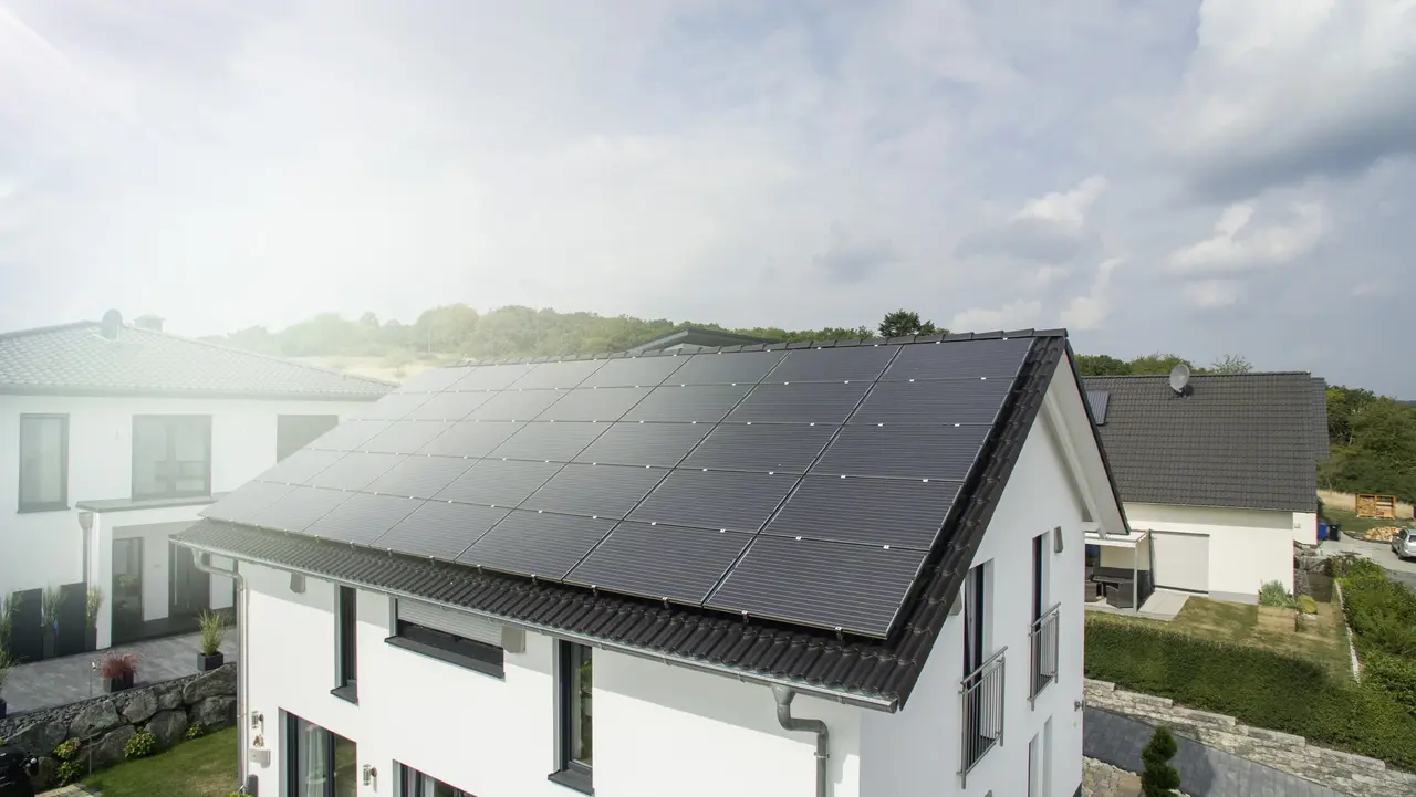  Modernes Einfamilienhaus mit großflächigen Solarpanelen auf dem Dach, sonniger Tag, benachbarte Häuser im Hintergrund.