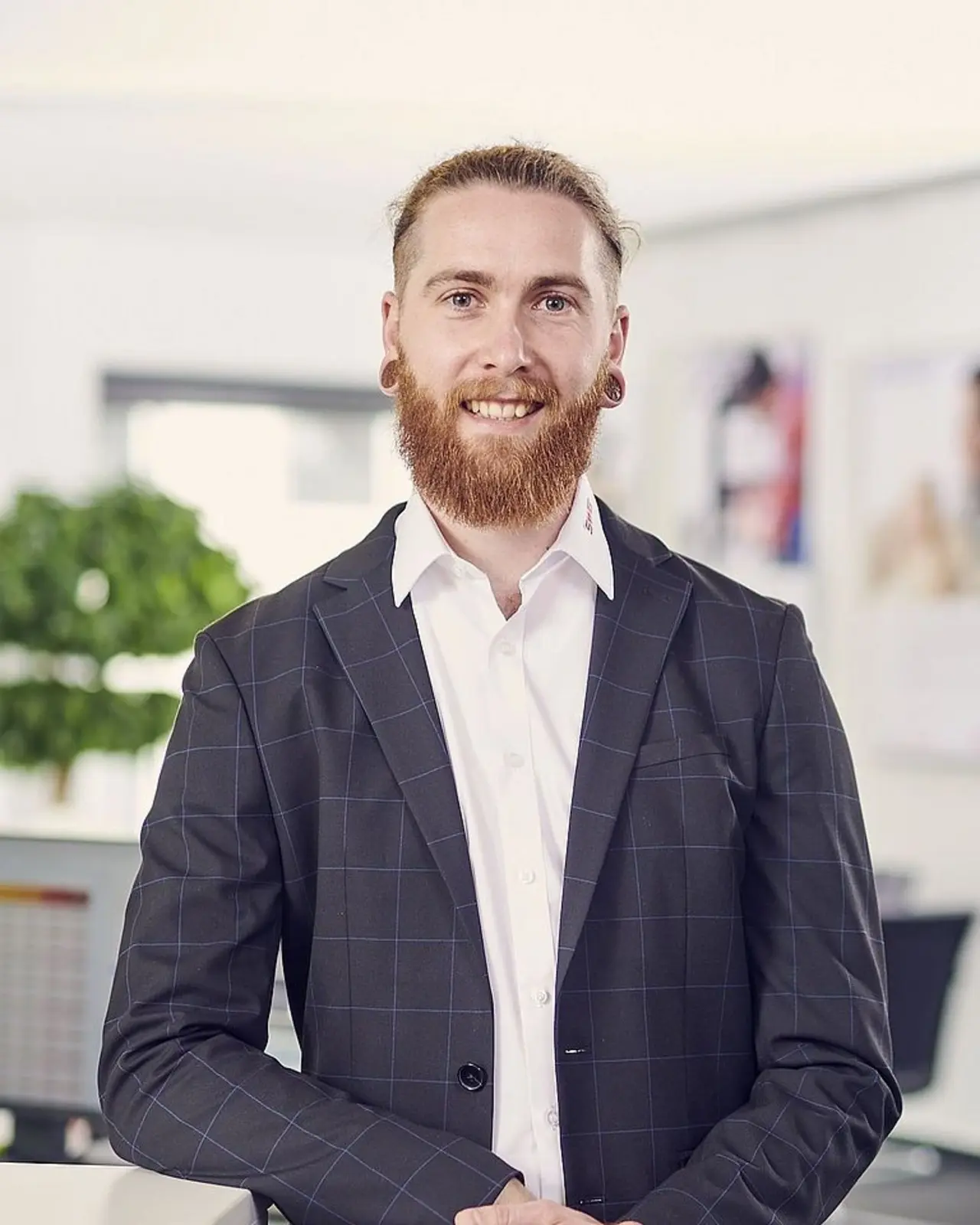 Ein Mann mit Bart und lockigem Haar steht in einem modernen Büro. Er trägt einen dunklen Anzug und ein weißes Hemd. Im Hintergrund sind verschwommen verschiedene Büroeinrichtungen und Pflanzen zu erkennen.
