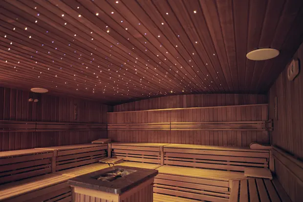 Auf dem Bild sieht man eine Sauna mit Saunabänken aus Holz mit einem eingebauten Sternenhimmel. In der Mitte des Raumes befindet sich das Herzstück der Sauna: Der Saunaofen.