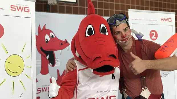 SWG-Maskottchen-Fabius-im-Trikot Das Bild zeigt ein rotes Drachenmaskottchen der Stadtwerke Gießen (SWG) in einem weißen Trikot mit der Aufschrift "SWG". Neben dem Maskottchen steht ein Mann mit einer roten Nase, Gesichtsbemalung und einem Taucherbrille, der ebenfalls den Daumen hoch zeigt. Beide Figuren lächeln und wirken fröhlich.
