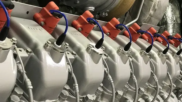 SWG-TREA2-BHKW-der-TREA-II Das Bild zeigt eine Nahaufnahme eines Teils eines industriellen Motors. Zu sehen sind mehrere zylinderförmige Bauteile, die in einer Reihe angeordnet sind. Die Oberseiten der Zylinder sind mit metallischen Abdeckungen versehen, auf denen elektrische Anschlüsse montiert sind, erkennbar an den blauen Kabeln, die von den orangefarbenen Steckverbindern abgehen.
