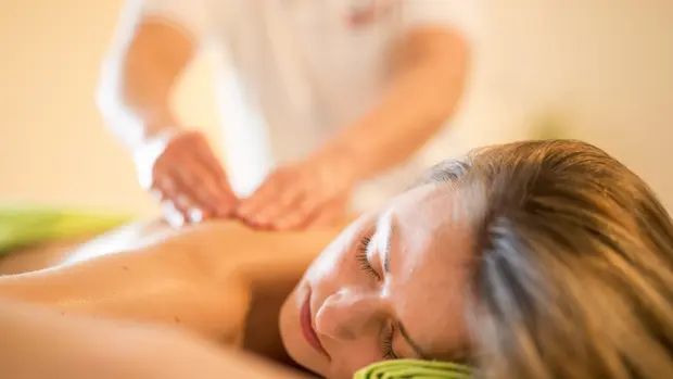 SWG-Massage-Hot-Stone Man sieht eine liegende Frau mit langen Haare, die von einem Masseur am Rücken massiert wird. Die Frau hat ihre Augen geschlossen und genießt die Massage.