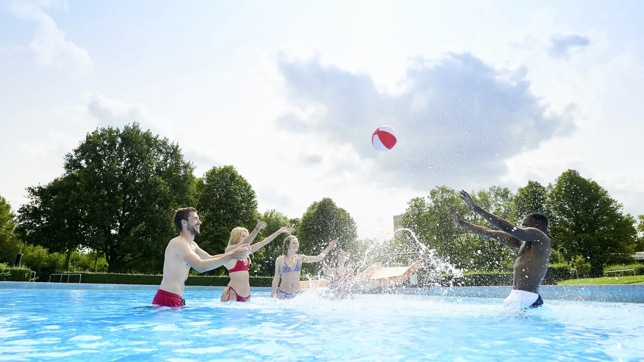 SWG-Menschen-spielen-Wasserball Auf dem Foto sieht man mehrere glückliche Menschen in einem flachen Becken Wasserball spielen. Der Ball befindet sich in der Luft und Wasser aus dem Becken spritzt nach oben.