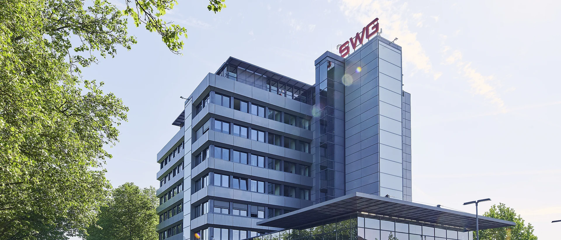 Ein modernes Bürogebäude mit Glas- und Metallfassade, markiert mit dem roten Logo "SWG" auf dem Dach. Umgeben von grünen Bäumen und unter klarem Himmel mit einigen Wolken.