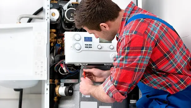 SWG-Heizungsmonteur Das Bild zeigt einen Handwerker, der an einer Heizungsanlage arbeitet. Er trägt ein rot-kariertes Hemd und blaue Arbeitshosen. Der Mann ist dabei, eine Reparatur oder Wartung an einer geöffneten Heiztherme durchzuführen. Die Anlage ist offen, sodass die inneren Komponenten, wie Rohre, Ventile und elektronische Steuerungen, sichtbar sind. Der Handwerker benutzt einen Schraubenzieher, um an der Anlage zu arbeiten, und konzentriert sich dabei auf seine Aufgabe.