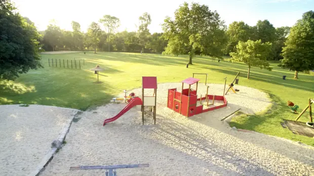 Das Foto wurde aus der Vogelperspektive aufgenommen und man erkennt einen Spielplatz mit einem Sandkastenbereich sowie eine Holzwippe, ein rotes Klettergerüst und ein rote Rutsche. Im Hintergrund sieht man weitläufige Rasenfläche, Bäume und die Sonne auf den Spielplatz scheinen.