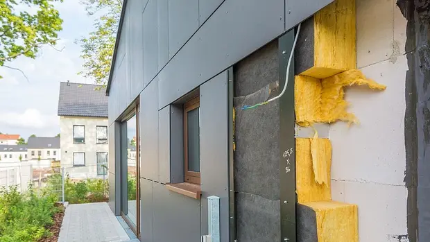 SWG-Dämmung-Haus s handelt sich um ein Foto von einer Gebäudefassade, bei dem ein Teil der Verkleidung entfernt wurde, um die darunterliegende Isolierung und Konstruktion zu zeigen. Man sieht Dämmmaterialien, wahrscheinlich Mineralwolle oder Glaswolle, die in die Wände eingebaut sind, um die Wärmeisolierung zu verbessern. Darüber hinaus sind einige Kabel zu sehen, die durch die Wand verlaufen.