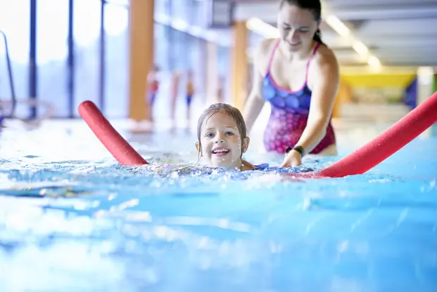 Dieses Bild zeigt ein kleines Mädchen beim Schwimmunterricht in einem Pool, unterstützt von einem Erwachsenen. Das Mädchen benutzt eine rote Poolnudel und lächelt. Im Hintergrund sieht man das gut beleuchtete Hallenbad und andere Personen.
