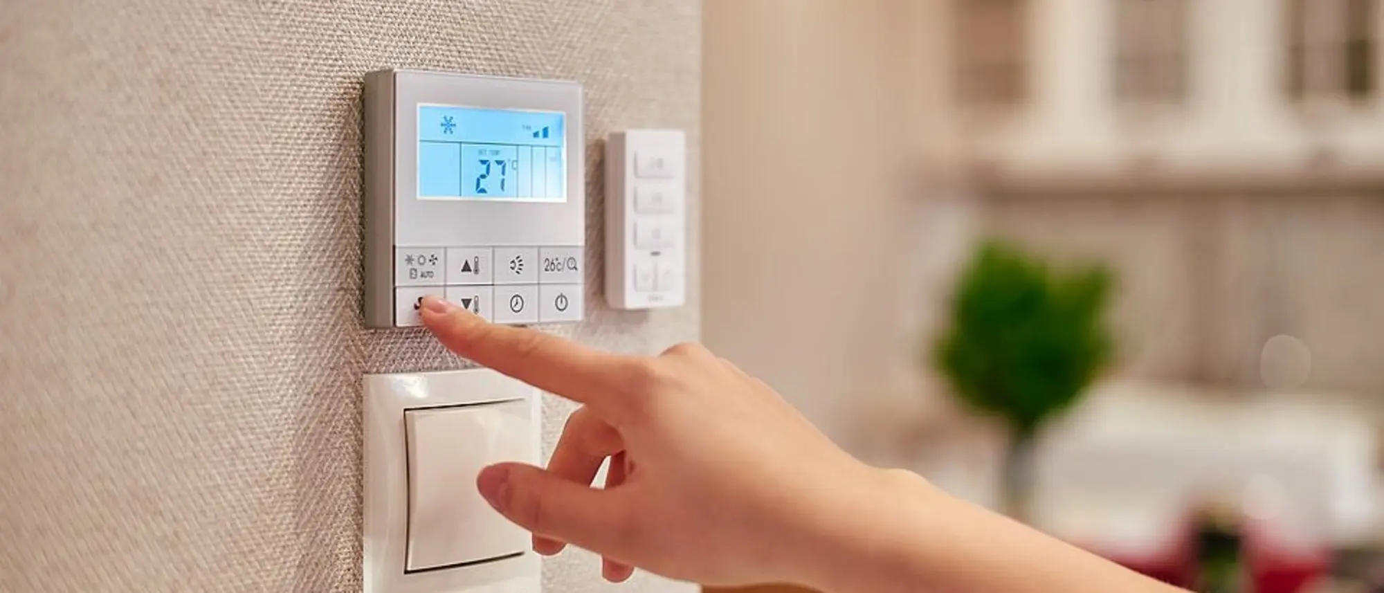 Das Bild zeigt eine Hand, die ein Wandthermostat bedient. Das Thermostat ist digital und zeigt auf dem Display eine Temperatur von 27°C an. Das Display ist beleuchtet, und es sind Symbole sichtbar, die auf verschiedene Einstellungen hinweisen, wie beispielsweise eine Schneeflocke, die für Kühl- oder Frostschutzfunktionen stehen könnte.