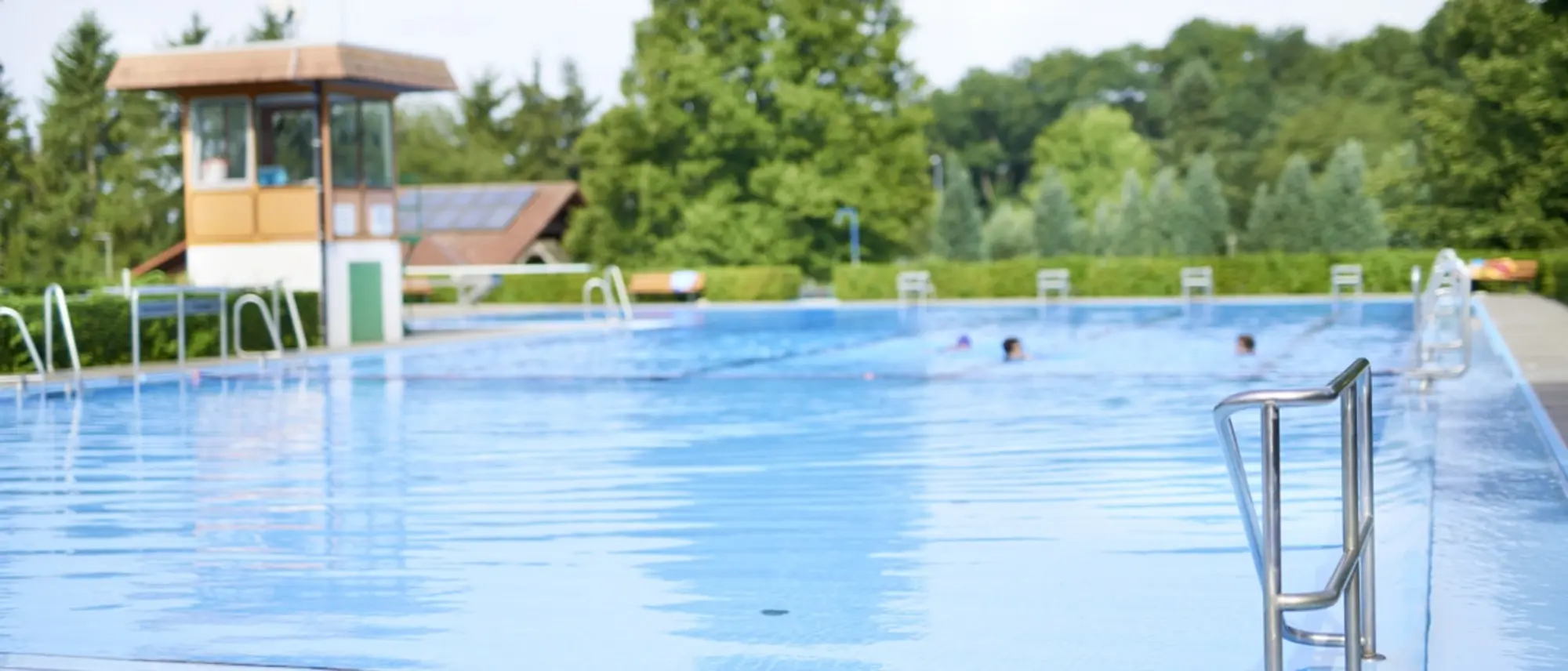 Man sieht den Einstieg mit Geländer in ein Freibadbecken mit mehreren Schimmern im hinteren Teil des Beckens.