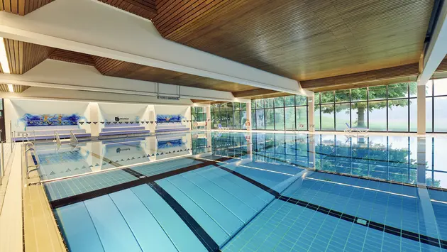 SWG-Westbad-Becken Ein modernes Hallenbad mit einem großen, hellblauen Schwimmbecken, das von Edelstahlgeländern umgeben ist. Die Decke besteht aus Holzpaneelen, die eine warme Atmosphäre schaffen. Im Hintergrund sind große Fenster, die viel Tageslicht hereinlassen und eine grüne Außenansicht bieten.