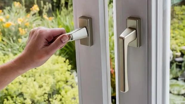 SWG-Fenstergriff Das Bild zeigt eine Nahaufnahme von zwei Fenstergriffen. Eine Hand greift gerade nach einem der Griffe und dreht diesen, vermutlich um das Fenster zu öffnen oder zu schließen. Die Griffe sind aus Metall, vermutlich in einer gebürsteten oder polierten Ausführung. Im Hintergrund sieht man einen Garten mit grünen Pflanzen und einigen gelben Blüten, was auf einen warmen, sonnigen Tag hinweist.