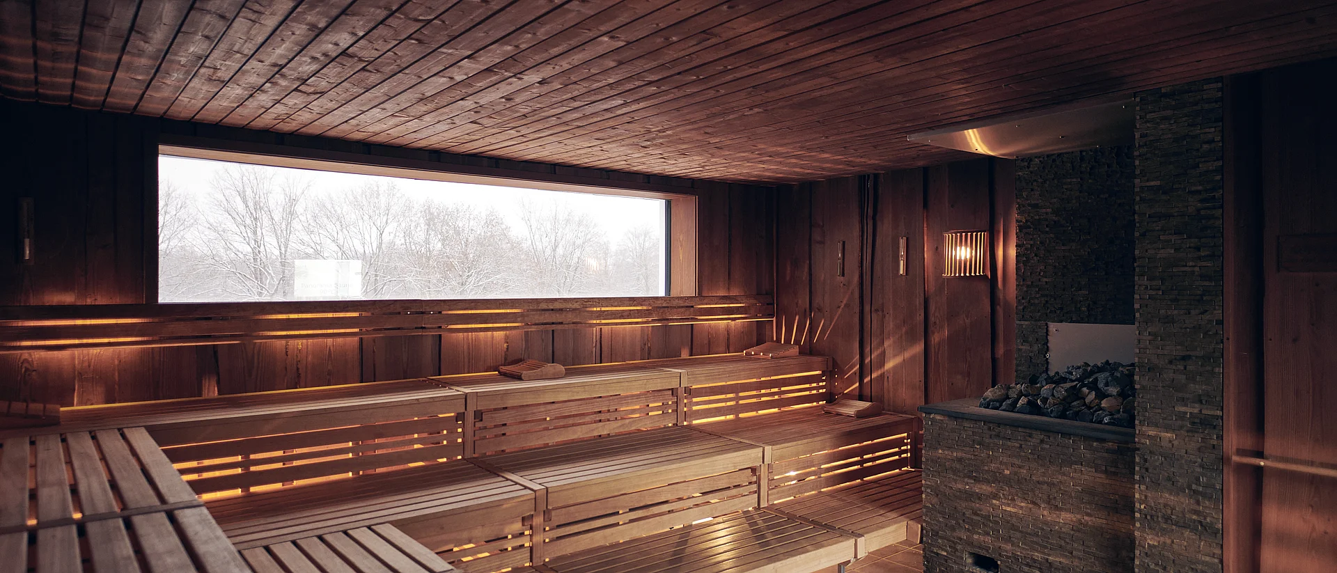 Auf dem Foto sieht man eine beleuchtet Sauna mit einem Fenster nach draußen. Auf der rechten Seite befindet sich er Saunaofen.