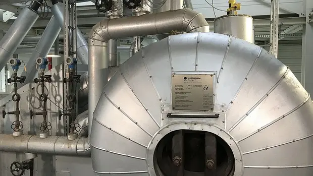 SWG_TREA2-Erzeugung-von-Dampf Das Bild zeigt eine industrielle Anlage mit einem großen, silbernen Gerät, das Teil eines Wärmetauschers ist. Das Gerät ist mit einer metallischen Isolierung ummantelt, um die Wärme effizient zu speichern und zu transportieren. Auf der Vorderseite des Geräts ist eine Platte mit technischen Spezifikationen und Sicherheitsinformationen angebracht.