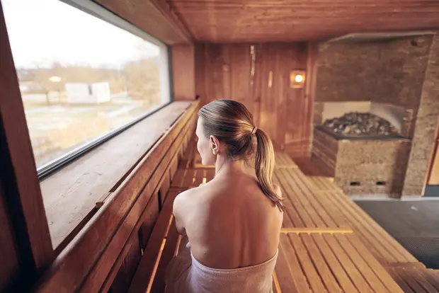 Auf dem Foto sieht man eine Frau mit Pferdeschwanz und einem Handtuch umgebunden in einer Sauna sitzen und aus dem Fenster schauen. Die Sauna besteht aus Holzbänken und auf der rechten Seite erkennt man den Saunaofen.