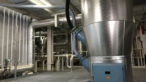 SWG-TREA2-Rauchgasreinigung Das Bild zeigt eine industrielle Anlage mit verschiedenen Rohrleitungen und Maschinenkomponenten, die mit Metall verkleidet sind. Im Vordergrund ist ein großer, trichterförmiger Metallbehälter zu sehen, der mit mehreren Schläuchen verbunden ist.