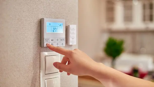 SWG-Heizungsregelung Das Bild zeigt eine Hand, die ein Wandthermostat bedient. Das Thermostat ist digital und zeigt auf dem Display eine Temperatur von 27°C an. Das Display ist beleuchtet, und es sind Symbole sichtbar, die auf verschiedene Einstellungen hinweisen, wie beispielsweise eine Schneeflocke, die für Kühl- oder Frostschutzfunktionen stehen könnte.