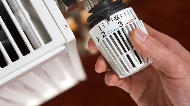 SWG-Thermostat Das Bild zeigt eine Nahaufnahme eines Heizkörperthermostats, das von einer Hand bedient wird. Das Thermostat befindet sich an einem Heizkörper, der traditionell in vielen Haushalten verwendet wird. Die Person stellt die Temperatur ein, indem sie das Thermostat dreht.